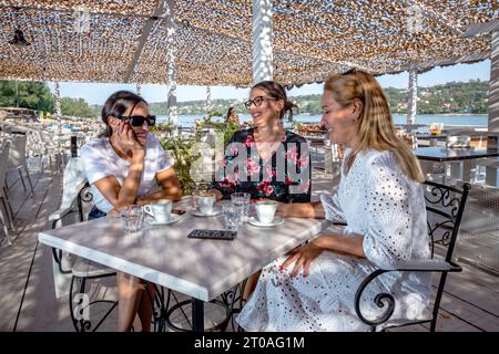 Drei fröhliche Freundinnen sitzen an einem Tisch, machen eine Kaffeepause, plaudern und lachen auf einer Restaurantterrasse am Fluss. Stockfoto