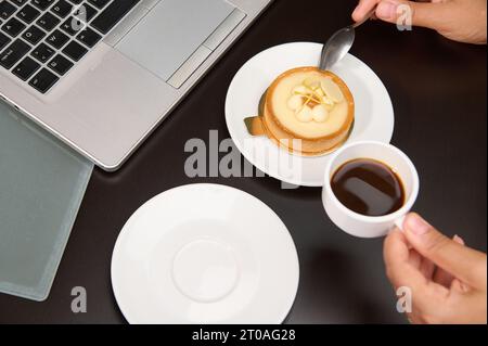 Eine Tasse Kaffee- und Zitronentörtchen auf dem Tisch mit teilweiser Sicht auf den Laptop. Konsumverhalten von Lebensmitteln und Getränken. Geschäftskonzept Stockfoto