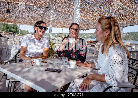Drei fröhliche Freundinnen sitzen an einem Tisch, machen eine Kaffeepause, plaudern und lachen auf einer Restaurantterrasse am Fluss. Stockfoto