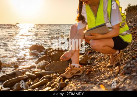 Nahaufnahme einer freiwilligen Frau, die eine Weste trägt und ein elektronisches Tablet hält, das den Boden am Strand analysiert. Im Hintergrund ist der Ozean, die Küste und der Sonnenuntergang. Konzept der Inspec Stockfoto