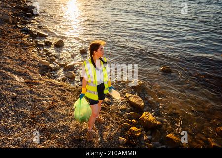 Sauber gemacht vom Kieselstrand. Draufsicht der jungen kaukasischen Frau, die Weste und Gummihandschuhe trägt, hält Plastiktüte mit Müll und Flaschenposierung. Konz Stockfoto