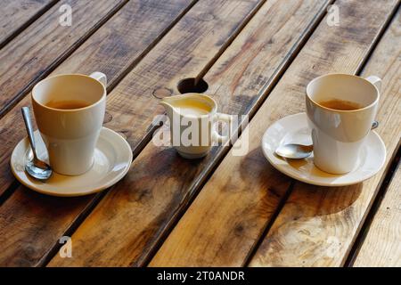 Zwei Tassen Kaffee und ein Milchkännchen auf einem Holztisch in einem Café im Freien. Stockfoto