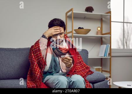 Ein kranker Mann mit Fieber sieht verwirrt auf ein Thermometer, das hohe Körpertemperatur anzeigt. Stockfoto