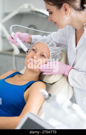 Junge Kosmetikerin in Gummihandschuhen tut Anti-Aging Verjüngung ohne chirurgische Behandlung für glückliche Frau in der medizinischen Praxis. Stockfoto