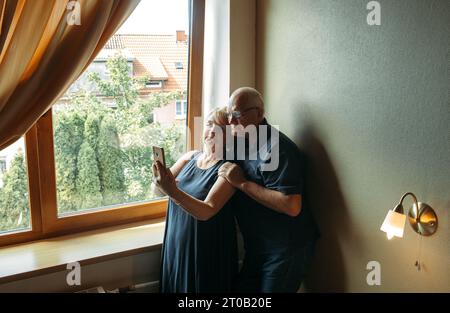 Ein glückliches älteres Paar steht am Fenster, posiert, macht ein Selfie am Telefon. Stockfoto