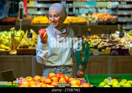 Porträt einer Käuferin in einem Supermarkt, eine muslimische Frau in einem Hijab wählt eine Apfelfrucht aus, glücklich mit einer großen Auswahl an Produkten und lächelt, Frau, die in einem großen Lebensmittelgeschäft einkauft. Stockfoto