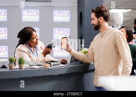 Glücklicher junger Mann, der am Checkout-Schalter im Bekleidungsgeschäft steht und einen mobilen Aktionscode verwendet, um den Black Friday Rabatt zu erhalten. Lächelnde afroamerikanerin, Kassierer, die Gutscheine auf dem Telefon des Kunden scannt Stockfoto