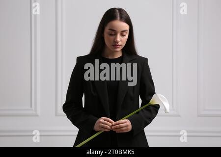 Traurige Frau mit Calla-Lilienblume in der Nähe der weißen Wand. Trauerfeier Stockfoto