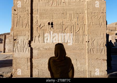 Stilvolle Frau, die eine braune Burka trägt, fängt einen Moment mit ihrem Handy ein und fotografiert die komplizierte Hieroglyphen auf KOM Ombo Stockfoto