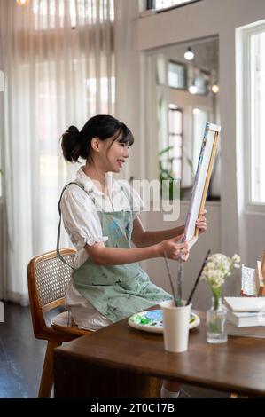 Ein Seitenbild einer schönen und talentierten jungen asiatischen Künstlerin auf einer Schürze mit Leinwand, die ihr gemaltes Bild in ihrem Minimalismus betrachtet Stockfoto