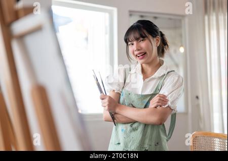Eine fröhliche und kreative junge asiatische Künstlerin in einer Schürze lächelt die Kamera an, steht mit überkreuzten Armen und hält Pinsel in der Hand Stockfoto