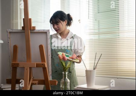 Eine nachdenkliche und kreative junge asiatische Malerin widmet sich in ihrem Atelier der Malerei auf einer Leinwand. Kreativität, Inspiration, Kunst-Worksho Stockfoto