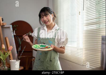 Ein Porträt einer positiven und kreativen jungen asiatischen Malerin auf einer Schürze, die einen Pinsel und ein Farbpalettengemälde auf einer Leinwand in h hält Stockfoto