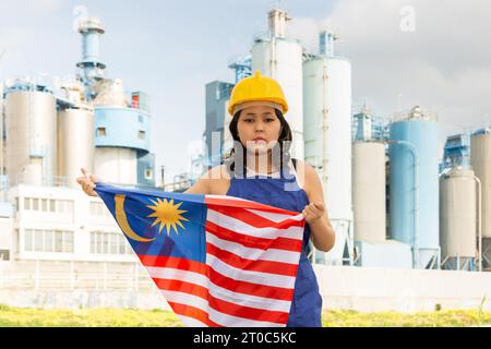 Besorgtes asiatisches Mädchen in Arbeitskleidung und Schutzhelm mit der Flagge malaysias vor der Industrielandschaft Stockfoto