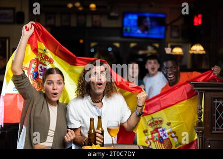 Begeisterte, vielfältige Sportfans mit spanischer Flagge, die ein Gewinnerspiel mit Gläsern Bier und Chips in der Kneipe feiern Stockfoto