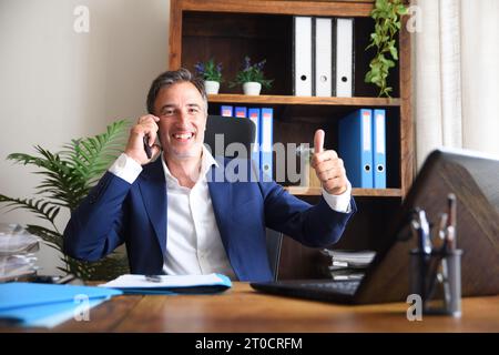 Ein lächelnder Geschäftsmann, der in seinem Büro auf dem Handy spricht, mit der Hand, die das OK-Schild mit Dokumenten und Laptop auf dem Tisch und Regalen zeigt Stockfoto