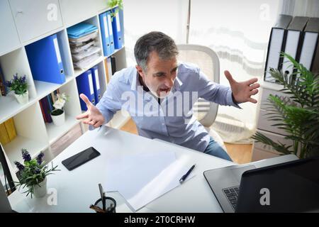 Wütender Mann vor einem Laptop, der auf einem Stuhl sitzt, in einem weißen Büro mit Regalen voller Aktenschränke, erhöhte Sicht Stockfoto