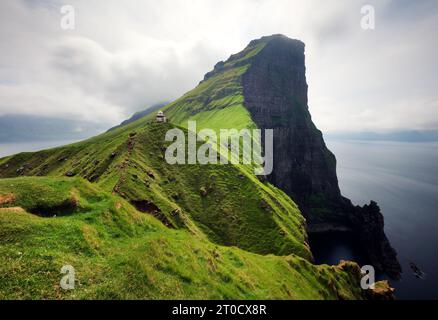 Der Leuchtturm auf einem grasbewachsenen Hügel in der Nähe einer felsigen schwarzen Klippe blickt an bewölkten Tagen über das ruhige Meer. Atemberaubender Blick aus der Luft auf ein kleines Stockfoto