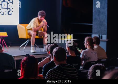 Verschwommener männlicher Trainer, der auf dem Stuhl auf der Bühne sitzt, in der Nähe des Bildschirms und während der Konferenz Fragen der Menge in der dunklen Halle beantwortet Stockfoto