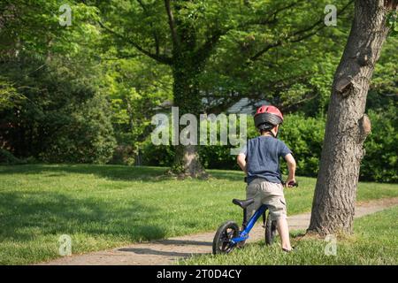 Kleiner Junge, der sich darauf vorbereitet, auf dem Bürgersteig in der Vorstadt mit dem Balance-Bike zu fahren. Stockfoto