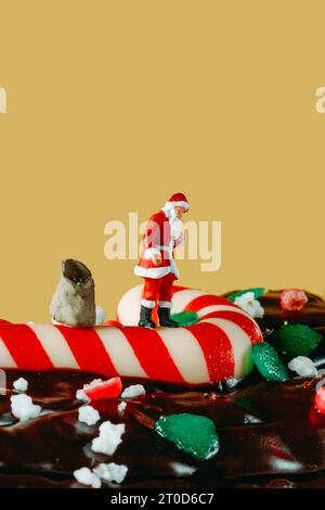 Ein Miniatur-weihnachtsmann steht auf einem Zuckerrohr neben einem Sack mit Geschenken, auf einem Baumkuchen, vor einem gelben Hintergrund mit etwas leerem Platz Stockfoto