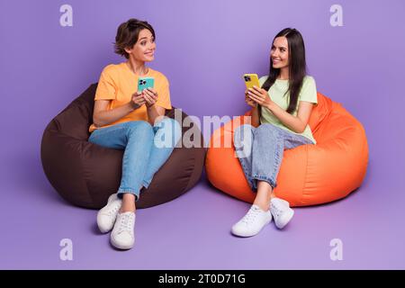 Ganzkörperfoto von Sit Sitzsack Freundschaft zwei Mädchen, die ihre iphones gemeinsam nutzen und isoliert auf violettem Hintergrund kommunizieren Stockfoto