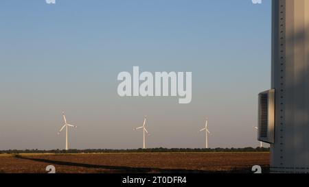 Windturbinen in einem in der Entfernung sichtbaren Feld vor dem Hintergrund eines klaren Abendhimmels und eines Fragments eines Kraftwerksturms im Vordergrund Stockfoto