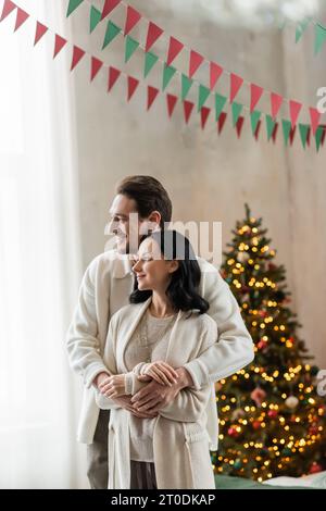 Glücklich verheiratete Paare zu Hause tragen sich umarmt und stehen zusammen neben dem verschwommenen Weihnachtsbaum Stockfoto