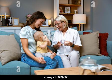 Großmutter, Mutter und Baby zu Hause zum Spielen, zum Spaß und zum Zusammenleben im Wohnzimmer. Liebe, glückliche Familie und Mutter, die ein Kind mit Gra trägt Stockfoto