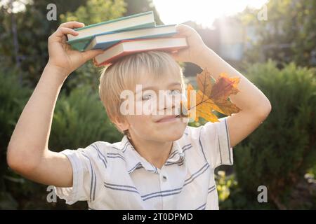 Der lustige blonde Junge hält ein orangefarbenes Herbstblatt im Mund und mehrere mehrfarbige Bücher liegen auf seinem Kopf. Zurück zur Schule, interessante Bildung, Spaß Stockfoto