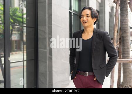 Junger asiatischer Geschäftsmann lächelt, während er zu seinem Arbeitsplatz vor modernen Firmengebäuden in der Stadt spaziert. Stockfoto