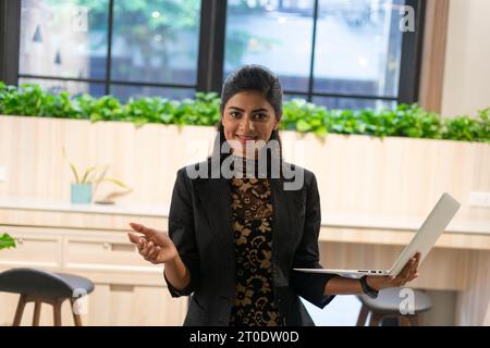 Glückliche junge asiatische Verkäuferin mit Laptop. Lächelnde Geschäftsfrau, Maklerin, im modernen Büro. Erfolgreiches Haltungsporträt. Stockfoto
