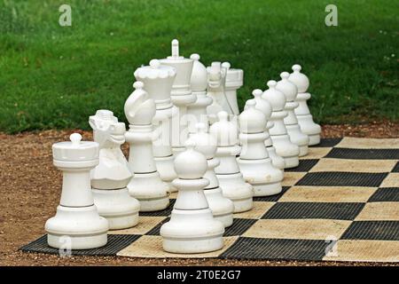 Riesige, weiße Schachfiguren auf einem Brett mit einem verschwommenen Hintergrund aus Sträuchern. Stockfoto