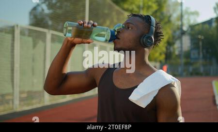 Müde afroamerikanischer Sportmann männlicher muskulöser Sportler Läufer Athlet Trinkwasser Sportflasche erfrischt Körperhydration nach dem Laufen auf Stadion Stockfoto