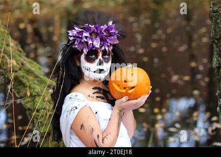 Ernsthaftes Mädchen im gruseligen Dead Bride Outfit mit Kürbis Jack o Laterne, die in die Kamera blickt, während sie während der Halloween-Feiertage in der Natur steht Stockfoto