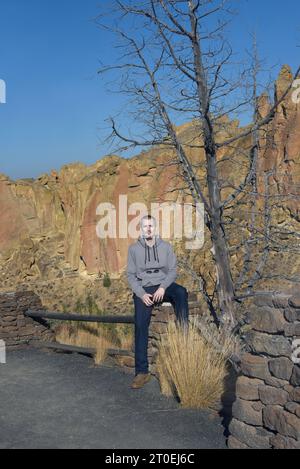 Besucher des Smith Rock State Park in Oregon ruhen auf einem Steinpfeiler auf dem Wanderweg. Kletterer sind im Hintergrund auf dem cl zu sehen Stockfoto