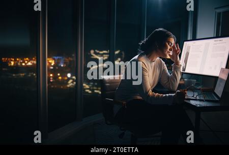 Erschöpfte kaukasische Geschäftsfrau, die nachts in einem schwach beleuchteten Büro arbeitet. Sie sitzt mit Laptop und Computer an ihrem Schreibtisch und hat Kopfschmerzen Stockfoto