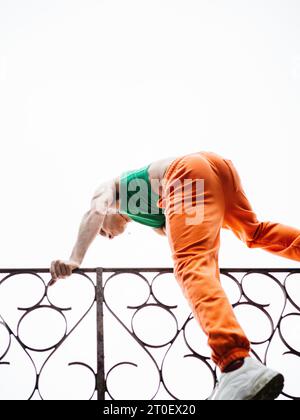 Nicht-binärer Mensch mit grünem Oberteil und orangefarbener Hose klettert über einen Zaun Stockfoto