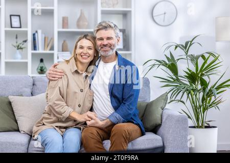 Porträt eines Reifen grauhaarigen Paares, eines glücklichen Familienmannes und einer glücklichen Frau, die lächelnd und in die Kamera blickend auf dem Sofa zu Hause sitzen. Stockfoto