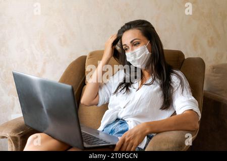 Niedliche Frau zu Hause im Stuhl mit Laptop mit Schutzmaske auf ihrem Gesicht kommuniziert auf einer Online-Konferenz, Konzept der Arbeit von zu Hause, Freiberufler oder Studium Stockfoto