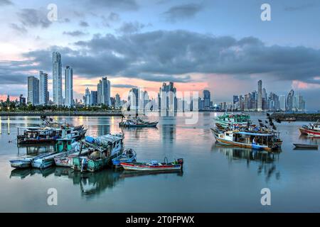 Futuristische Skyline der Stadt Panama bei Sonnenuntergang mit Fischerbooten im Vordergrund, die sich im stillen Wasser spiegeln. Stockfoto