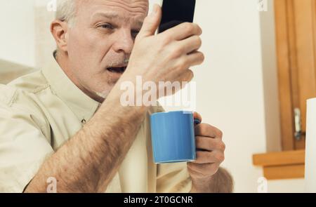 Frühstückszeit: Ein älterer Mann in der Küche schielt auf sein Telefon. Moderne Designs sind nichts für Sehbehinderte. Ein Augenarzt-Besuch und sp Stockfoto