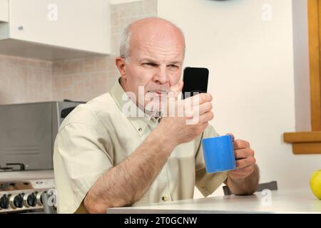 Ein älterer Mann in der Küche schielt während des Frühstücks auf sein Smartphone. Bei der heutigen Gestaltung von Telefonen werden Sehbehinderte möglicherweise vernachlässigt. Ein Besuch beim Optometristen Stockfoto