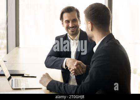 Glückliche junge Geschäftsleute in formellen Jacken, die am Arbeitsplatz die Hand schütteln Stockfoto