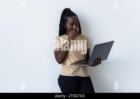 Überraschtes, schockiertes afroamerikanisches Mädchen, isoliert auf grauem Studiohintergrund, in einem weißen Pullover, hält einen Laptop in der Hand und schaut ihn an, Cover Stockfoto