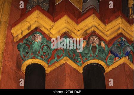 Geschnitzte Kapitelle auf der linken Seite des Hauptportals der mittelalterlichen Kathedrale St. Georg in Limburg-an-der-Lahn, Hessen, Deutschland. Ein Vogel pickt auf einen Haufen blauer Trauben (rechts) und Gesichter blicken aus grünem Laub hervor, vor einem Hintergrund von lebhaftem Orange-Rot und Gelb. Diese spätromanische/frühgotische Kathedrale, die in den späten 1100er/frühen 1200er Jahren n. Chr. erbaut wurde, erhielt durch Restaurierungsarbeiten in den 1960er und 70er Jahren ihr ursprüngliches überschwängliches und farbenfrohes Aussehen zurück, wobei die Farben durch Spuren der Originalfarbe bestimmt wurden. Stockfoto