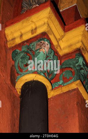 Skulpturenhauptstadt auf der Nordseite des Hauptportals der mittelalterlichen Kathedrale St. Georg in Limburg-an-der-Lahn, Hessen, Deutschland. Gesicht umgeben von grünen Blättern, vor einem Hintergrund von lebhaftem orange-rot und gelb. Die spätromanische/frühgotische Kathedrale St. Georg, die in den späten 1100er/frühen 1200er Jahren n. Chr. erbaut wurde, erhielt durch Restaurierungsarbeiten in den 1960er und 70er Jahren ihr ursprüngliches überschwängliches und farbenfrohes Aussehen zurück, wobei die Farben durch Spuren der Originalfarbe bestimmt wurden. Stockfoto