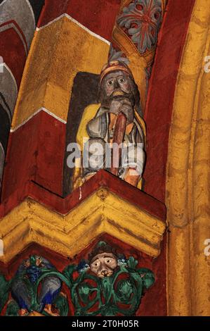 Die Figur des Heiligen Nikolaus befindet sich über den Kapitellen auf der linken Seite des Hauptportals des mittelalterlichen St. Georg-Doms in Limburg-an-der-Lahn, Hessen. Auf den Kapitellen ist ein Vogel abgebildet, der an einem Haufen blauer Trauben pickt (links) und ein menschliches Gesicht, umgeben von grünem Laub, vor einem Hintergrund von lebhaftem orange-rot und gelb. Diese spätromanische/frühgotische Kathedrale, die in den späten 1100er/frühen 1200er Jahren n. Chr. erbaut wurde, erhielt durch Restaurierungsarbeiten in den 1960er und 70er Jahren ihr ursprüngliches überschwängliches und farbenfrohes Aussehen zurück, wobei die Farben durch Spuren der Originalfarbe bestimmt wurden. Stockfoto