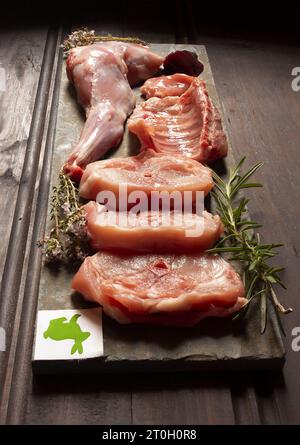 Geschnittenes weißes Kaninchenfleisch mit Thymian- und Rosmarin-Zweigen auf einem Schieferbrett auf schwarzem Hintergrund. Fleischindustrie. Es hat ein Etikett von einem Kaninchen in Stockfoto