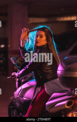 Fotografie mit blauen und rosa Neonfarben auf einem Motorrad von hinten. Porträt eines jungen blonden kaukasischen Models mit schwarzer Lederjacke Stockfoto